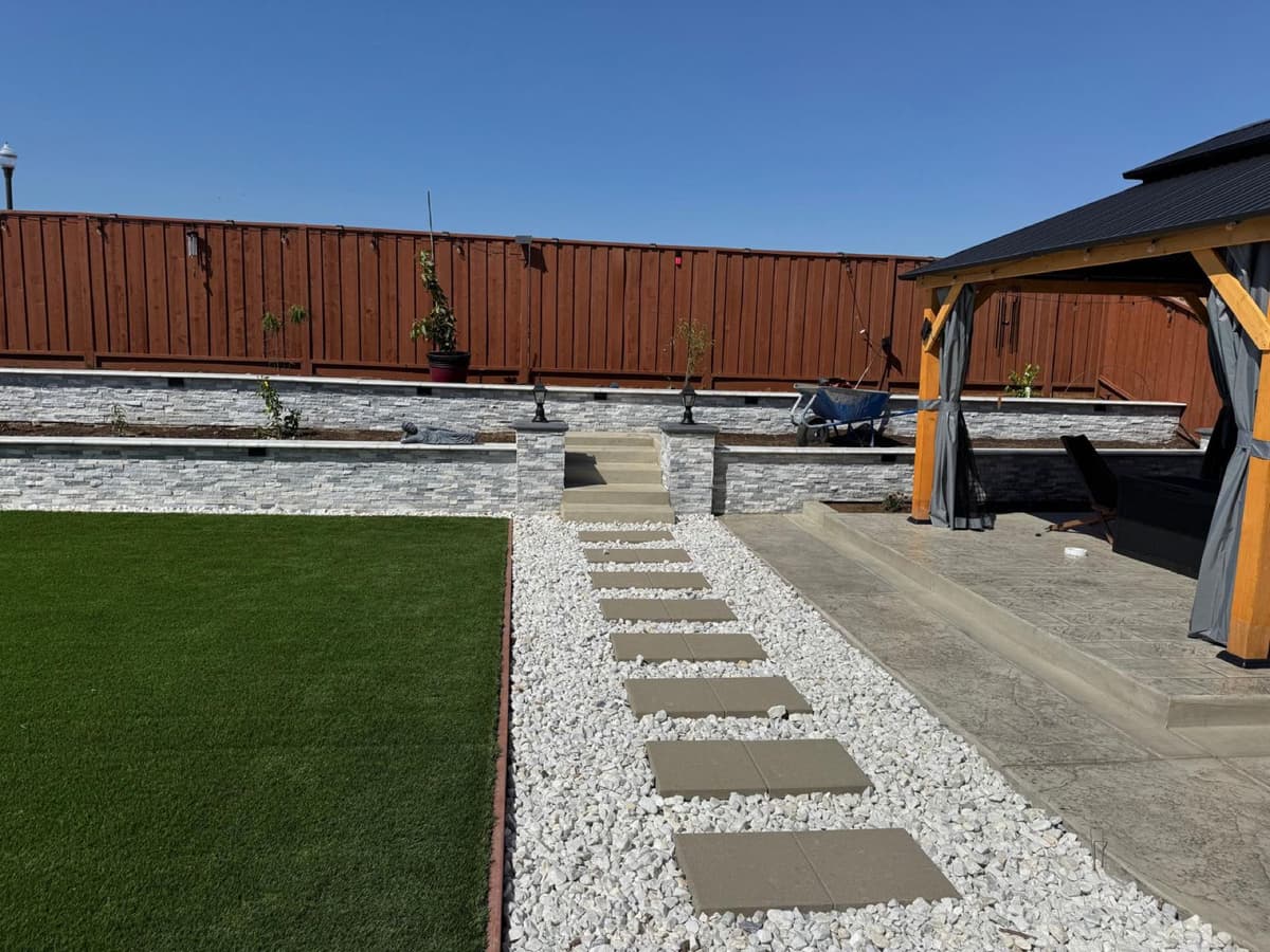 backyard with stacked stone retaining walls, turf lawn, gravel stepping path, and a pergola sitting area in Stockton, California